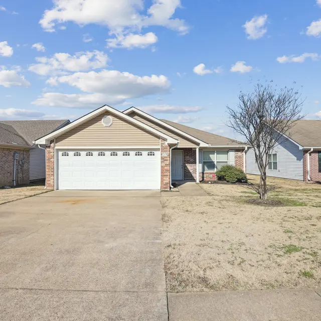 Suburban House Front View A single-story suburban house with a light brown exterior and a two-car garage. The driveway is paved, and there is a small tree in the yard. The sky is partly cloudy with blue patches visible.
