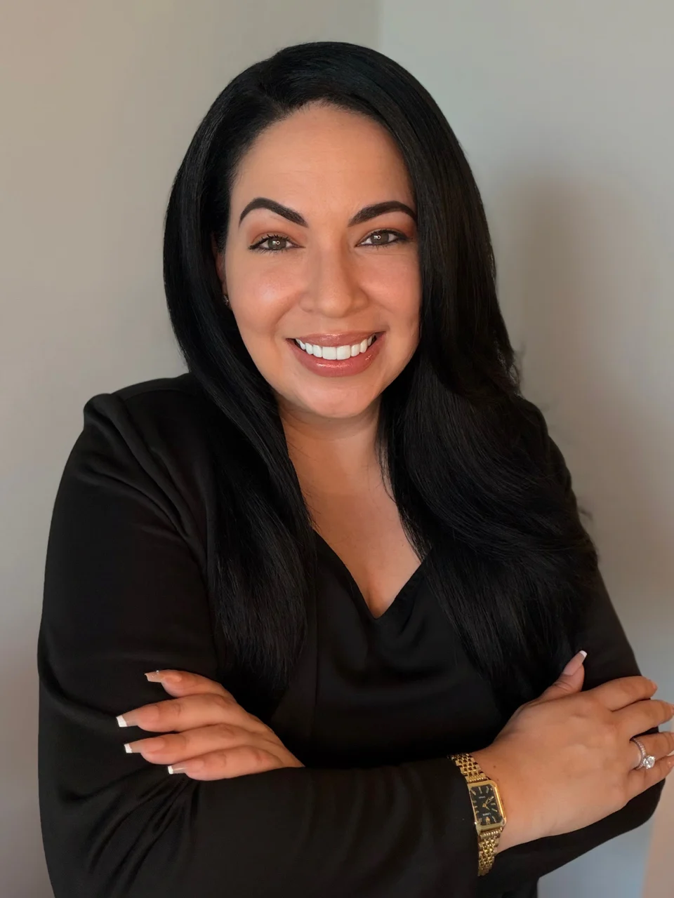 A smiling woman with long dark hair, wearing a black blouse, crossing her arms confidently against a light gray background.