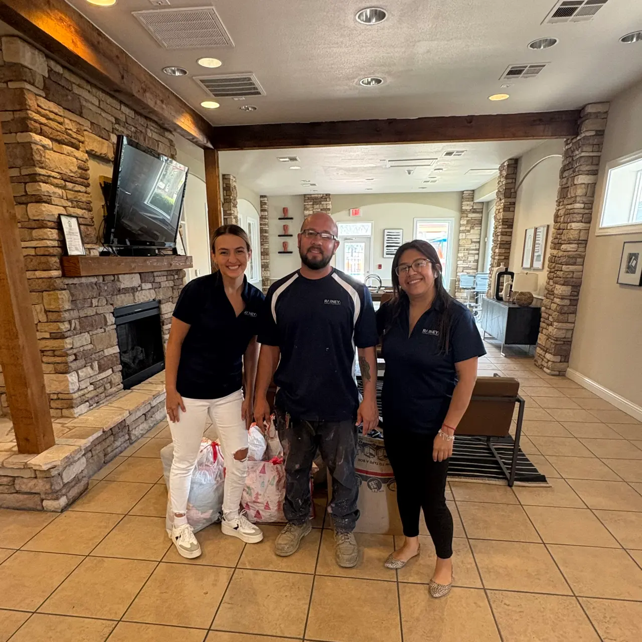 Three individuals posing together in a reception area with stone walls and a fireplace, smiling at the camera. Two women are wearing dark shirts and light-colored pants, while a man in the center is wearing a dark shirt and glasses. They are holding bags of items.