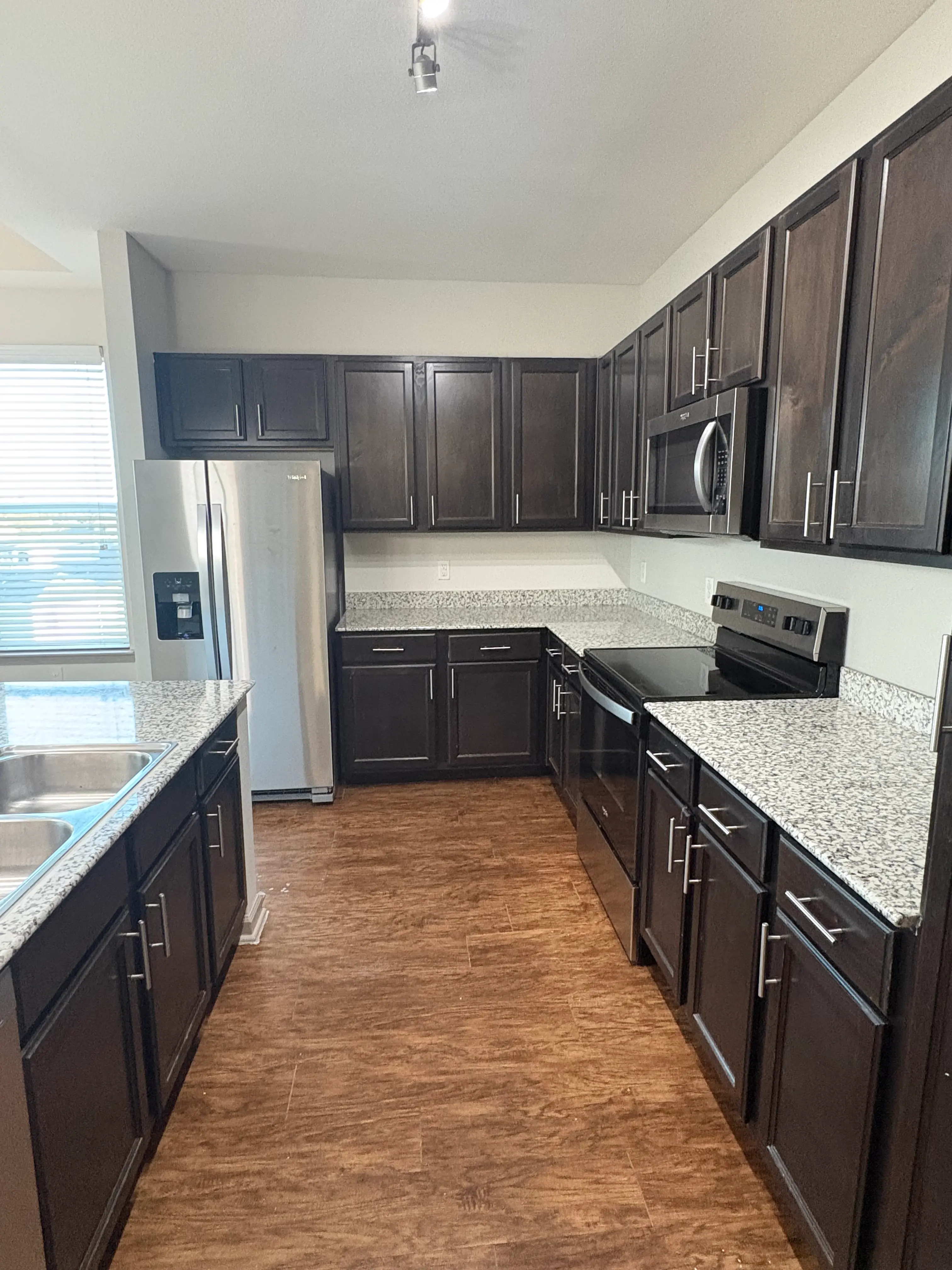 A modern kitchen featuring dark wooden cabinets, granite countertops, stainless steel appliances, and an open layout with a window letting in natural light.