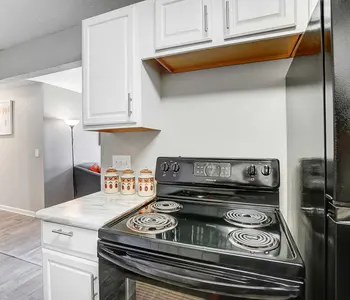 A modern kitchen featuring a black electric stove, white cabinetry, and a countertop. The background includes a glimpse of a living area with a lamp and a wall art piece.
