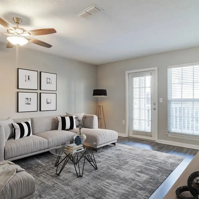 A modern living room with a light-colored sofa, decorative pillows, a coffee table, and a rug on the floor. The room features a ceiling fan, artwork on the walls, and sliding glass doors leading to a balcony with blinds.