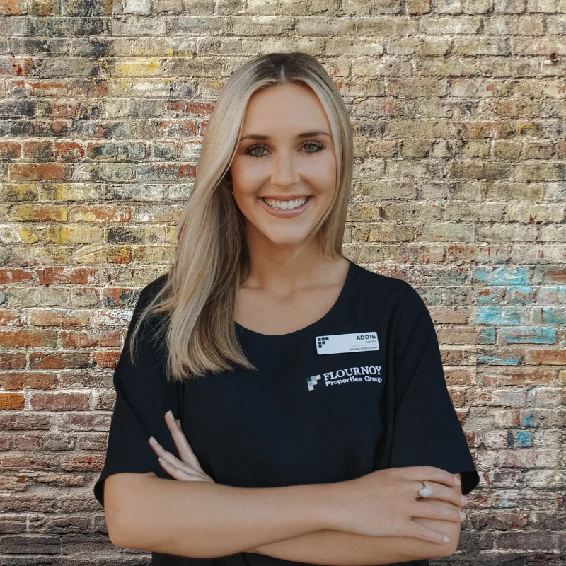 A smiling woman with long blonde hair stands in front of a textured brick wall. She wears a black shirt with the text 'FLOURNOY Property Group' visible. Her arms are crossed, and she appears confident and approachable.