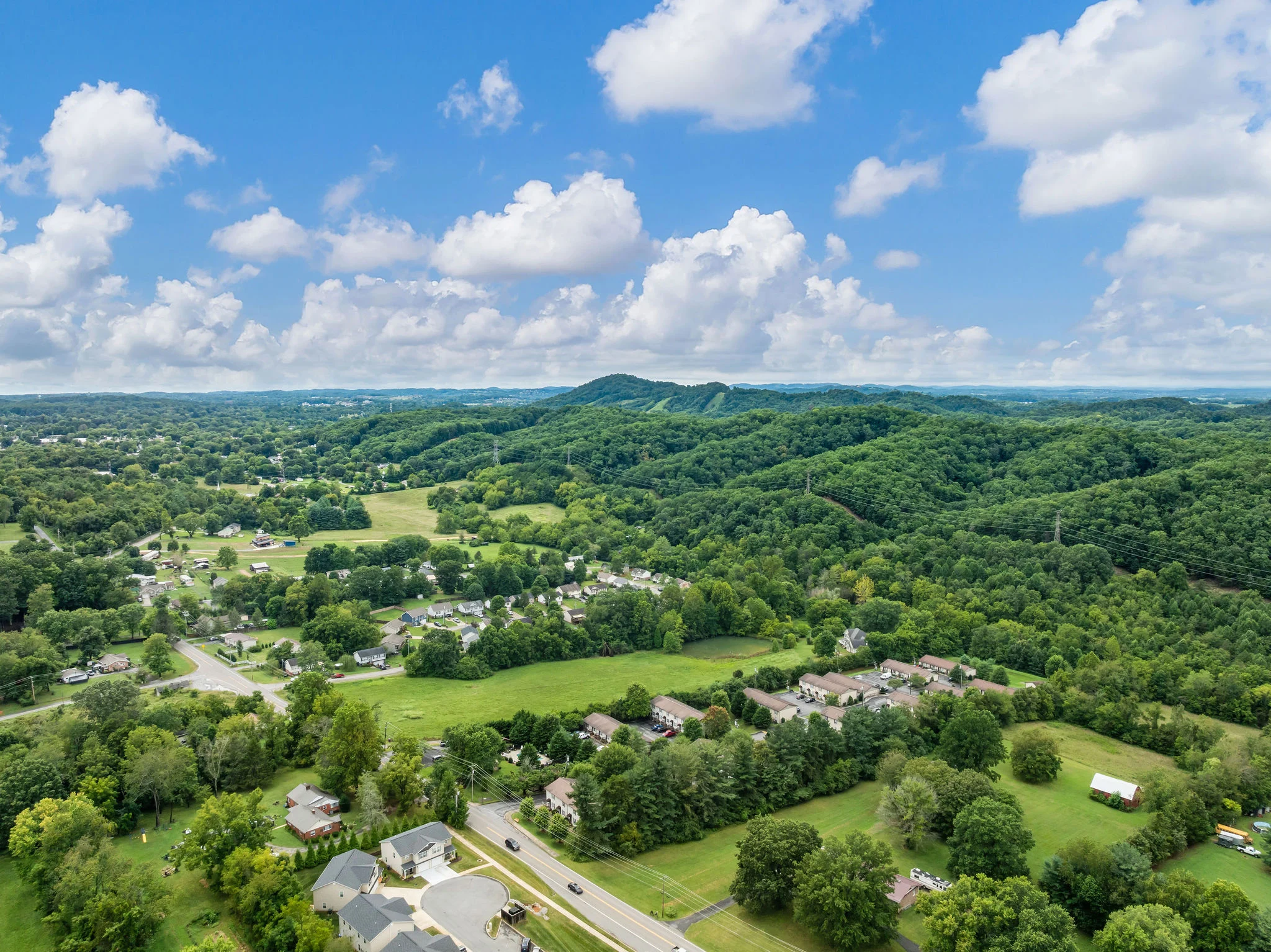 Aerial View of Green Hills and Homes Aerial view of lush green hills and residential areas under a bright sky with scattered clouds.