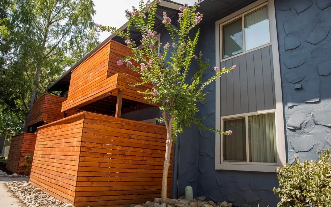 A modern exterior view of a house featuring wooden structures and a tree, with a textured blue wall and green surroundings.