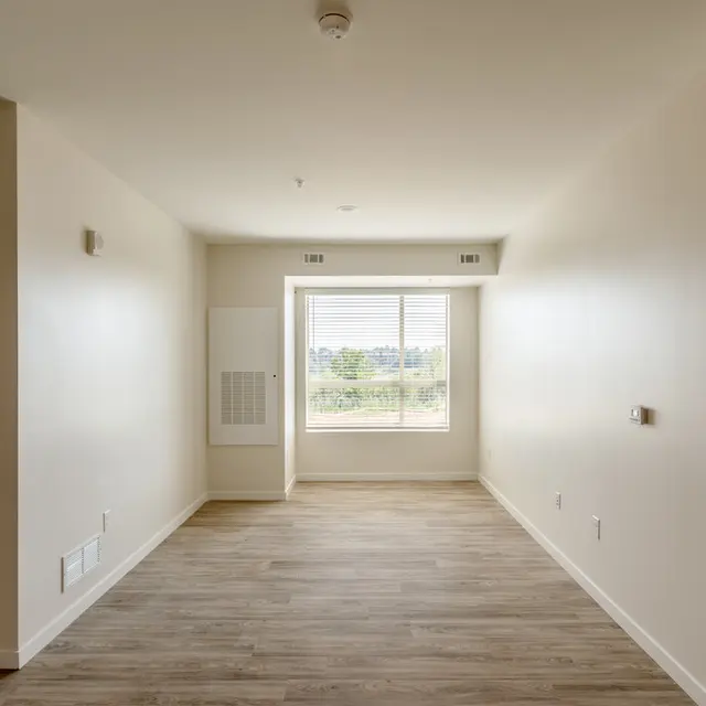 Empty interior of an apartment with beige walls and wood-like flooring, featuring a large window.