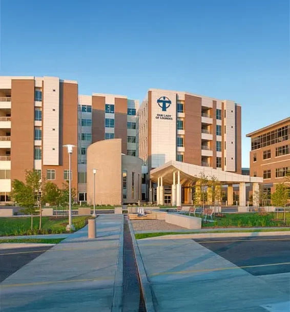 An exterior view of a modern hospital building featuring several stories, large windows, and a welcoming entrance area with trees and landscaped gardens.