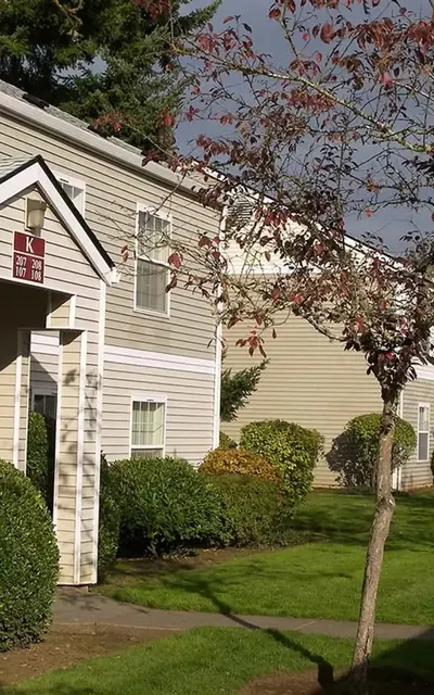 A view of an apartment complex with beige siding and a green lawn, featuring trees and a walkway.