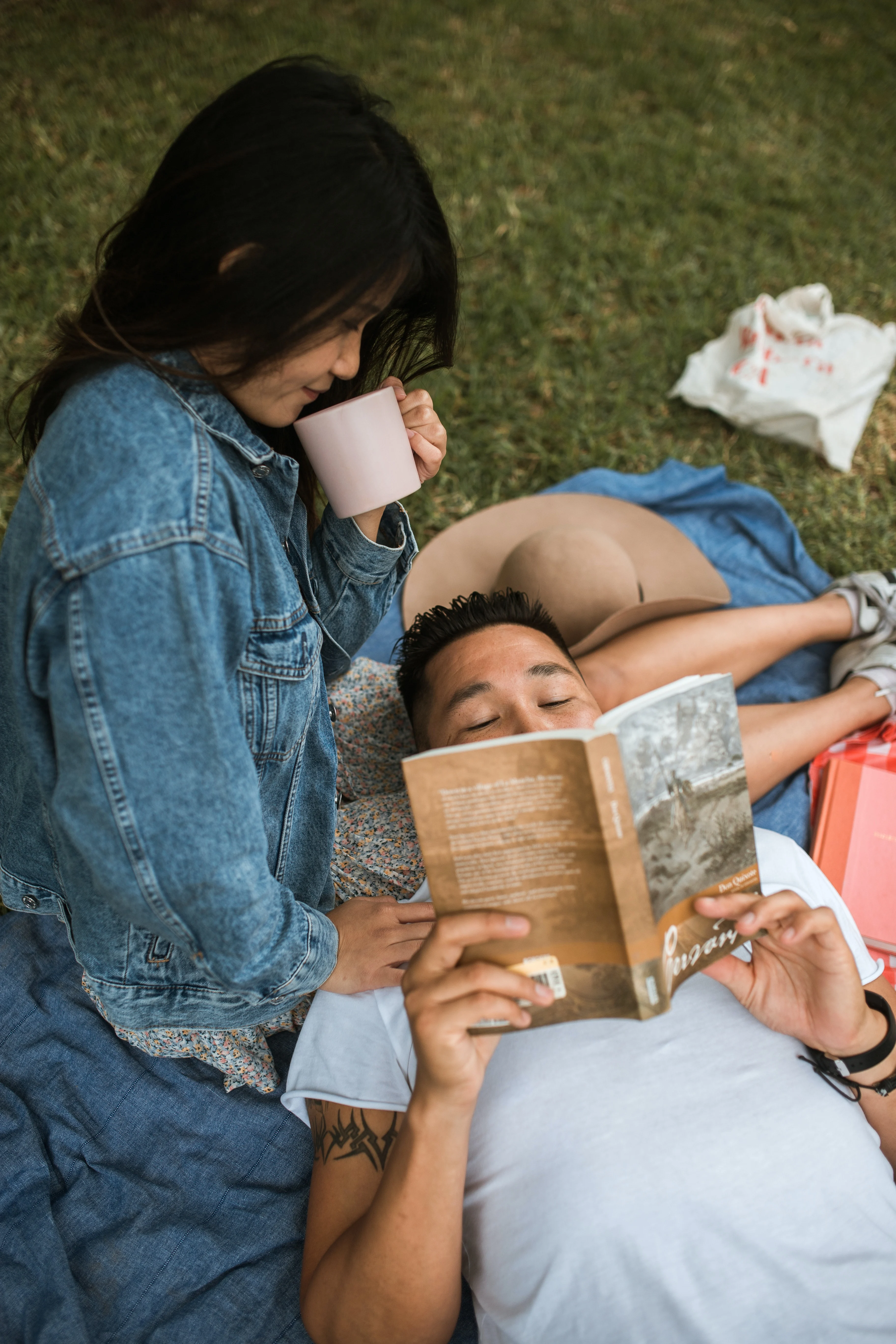 A couple lounging on a blanket in a grassy area. One person is lying on their back reading a book while the other is standing beside them holding a mug. They both appear relaxed and comfortable in a casual outdoor setting.