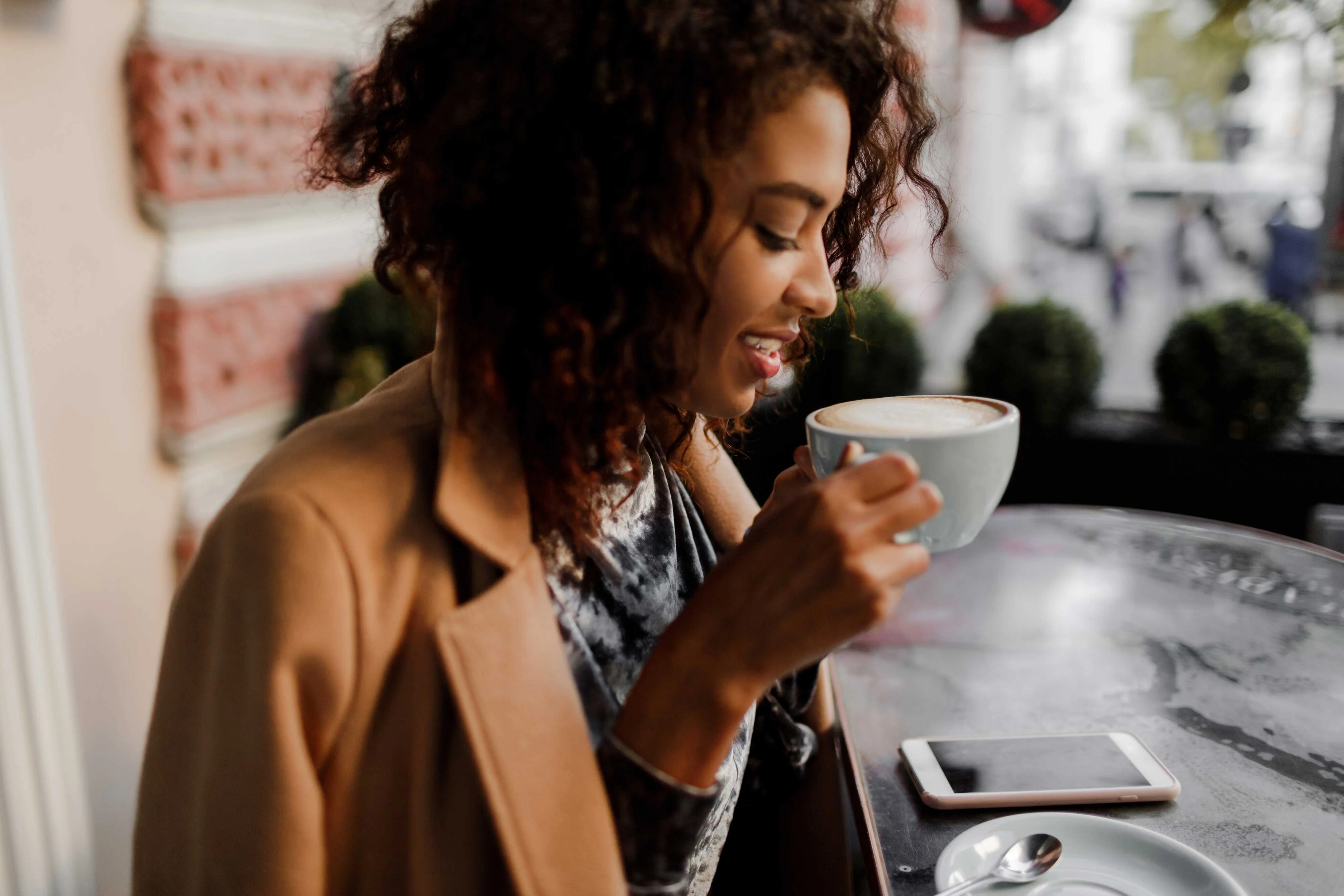 A woman with curly hair enjoys a cup of coffee at a café, looking down at her drink with a smile. A smartphone is placed on the table beside her.
