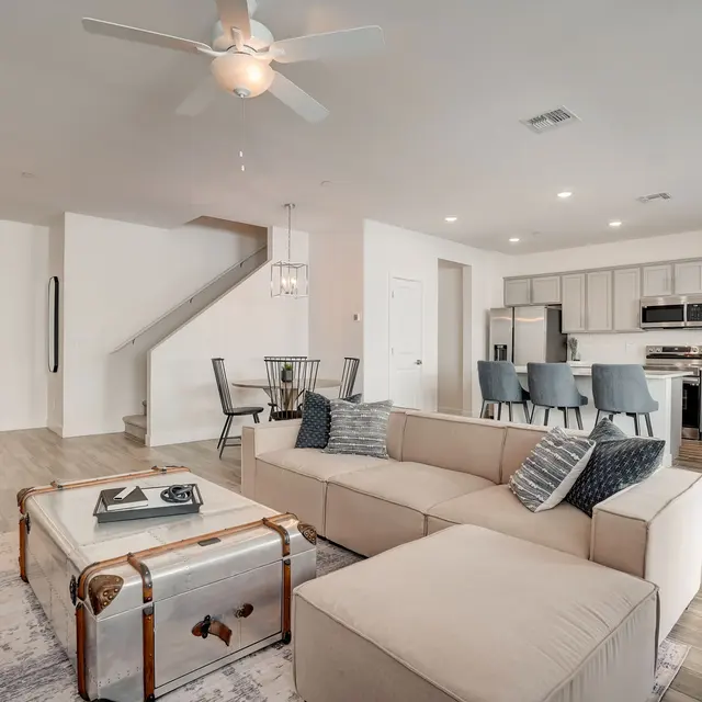 Contemporary Living Room with Open Layout A modern living room featuring a large sectional sofa, a coffee table with a vintage design, a dining area with black chairs, and a staircase leading to the upper floor. The decor is light and inviting, with wooden flooring and soft lighting.