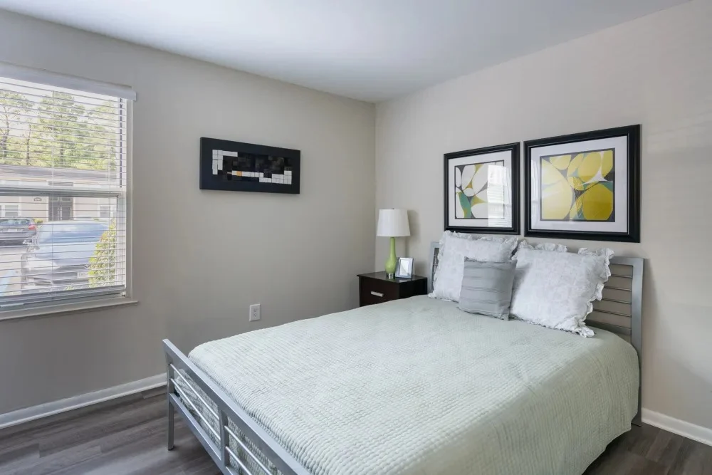 Cozy Bedroom Interior A cozy bedroom featuring a double bed with a light green cover, two framed artworks on the wall, a bedside lamp, and a window with blinds letting in natural light.