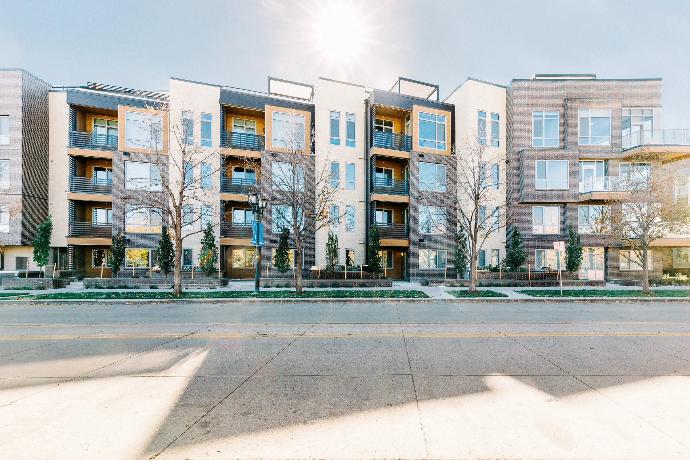 A contemporary apartment building featuring several balconies and large windows, with a sidewalk in the foreground and trees lining the property.
