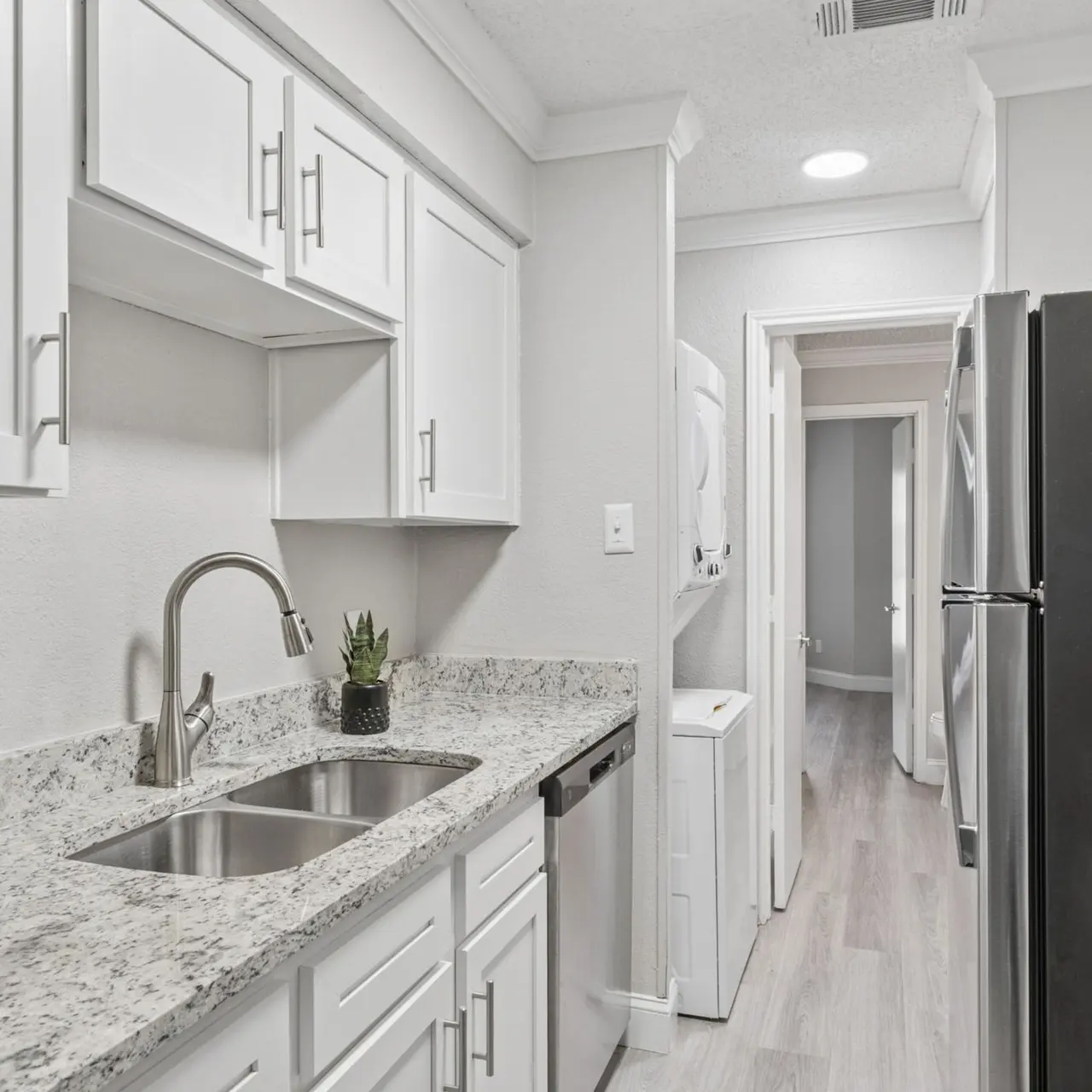 A modern kitchen with white cabinets, granite countertops, and stainless steel appliances. The kitchen includes a double sink and a black refrigerator, with a glimpse of a laundry area in the background.