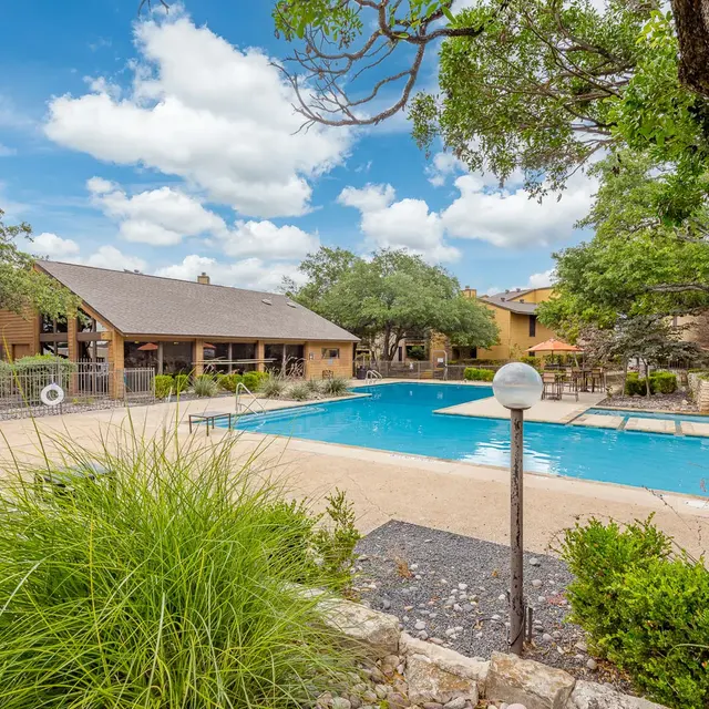 A serene community pool area with a large swimming pool surrounded by greenery and trees, featuring a clubhouse in the background under a blue sky with fluffy clouds.