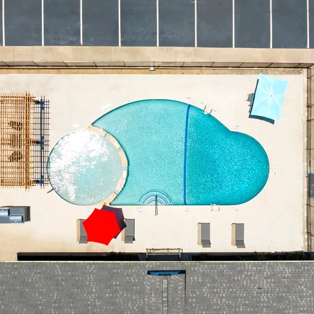 Aerial view of a swimming pool shaped like a raindrop, surrounded by a wooden deck, lounge chairs, and umbrellas. Parking spaces are visible in the background.