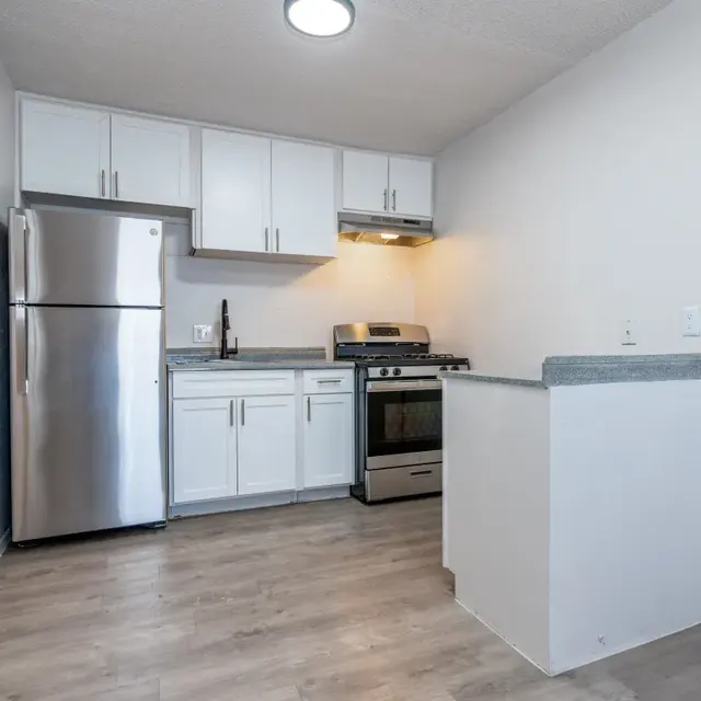 A modern kitchen with white cabinets, stainless steel appliances, and wooden flooring. The kitchen features a refrigerator, stove, and a small bar area.