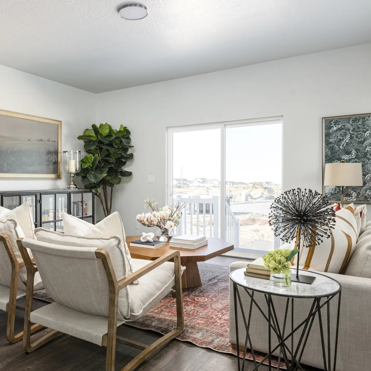 A modern living room featuring a white sofa, two wooden armchairs, a coffee table, and large windows letting in natural light. A potted plant adds greenery, and decorative art pieces adorn the walls.