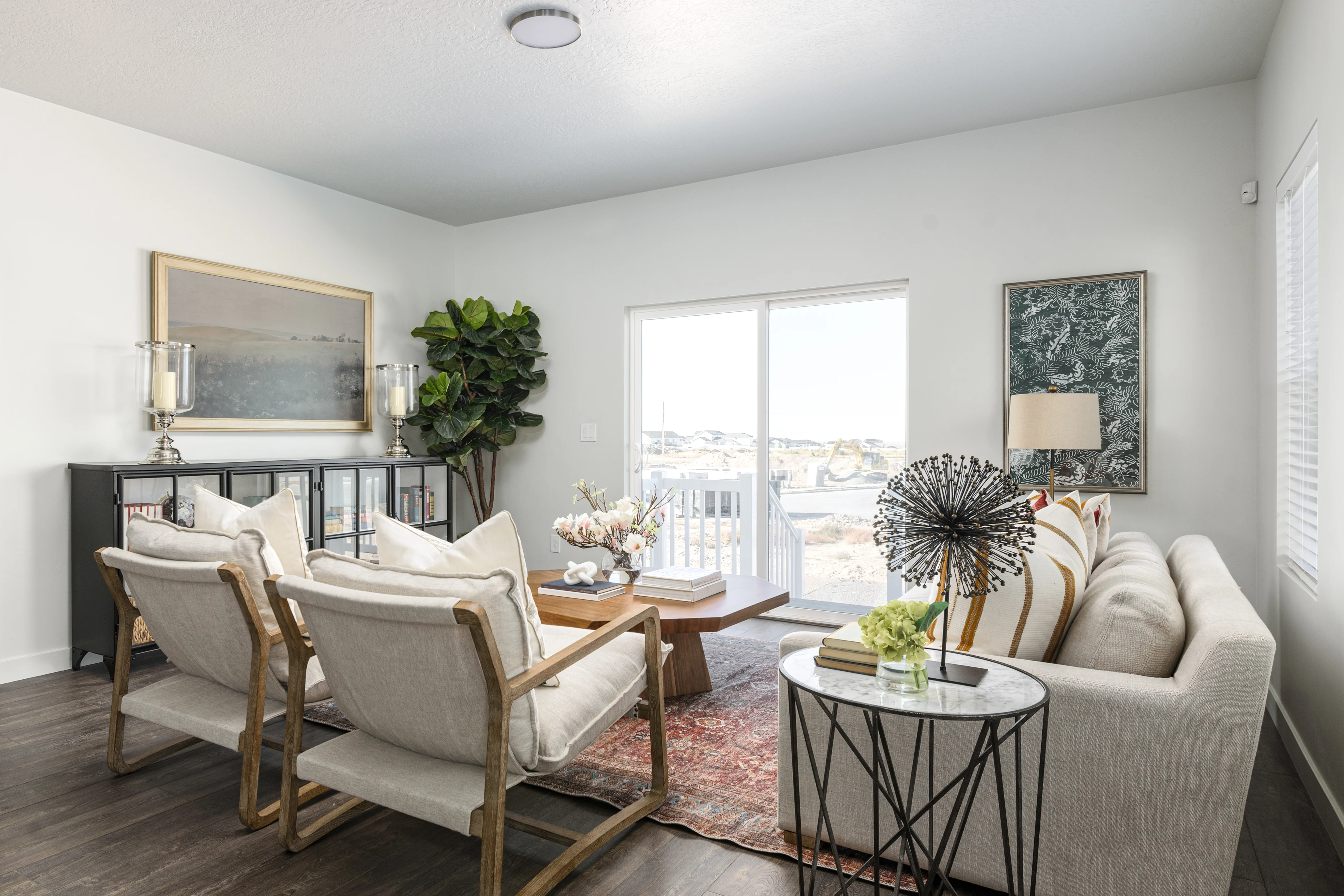 A modern living room featuring a white sofa, two wooden armchairs, a coffee table, and large windows letting in natural light. A potted plant adds greenery, and decorative art pieces adorn the walls.