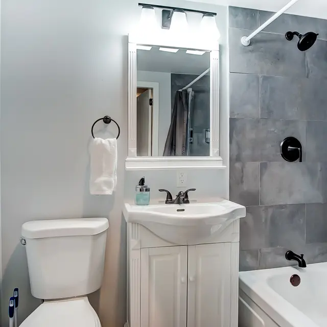 A clean and modern bathroom featuring a white vanity, toilet, and a bathtub with gray tiled walls.