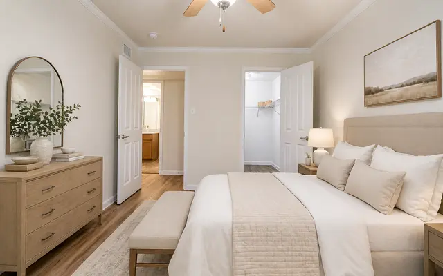 A well-decorated bedroom featuring a queen-sized bed with beige bedding, a small bench at the foot of the bed, wooden nightstands with lamps, and a large dresser with decorative items. The room has a ceiling fan and a neutral color palette, with an open closet visible through a doorway.