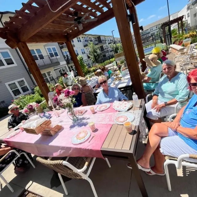 A group of people sitting around a table outdoors under a wooden structure, enjoying a meal together at the Best Garden Competition Event. The table is set with plates and decorations, with people smiling and chatting in a sunny environment.