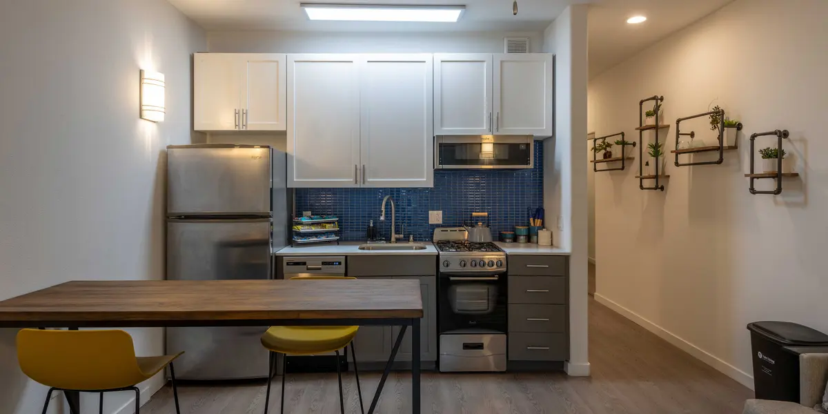 A modern kitchen featuring white cabinetry, stainless steel appliances, a blue tiled backsplash, and a dining area with a wooden table and yellow chairs.