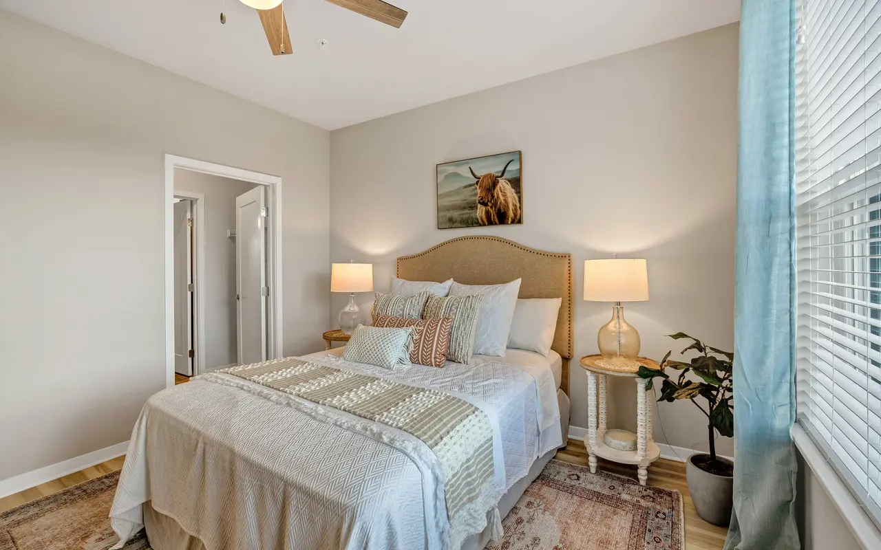 A cozy modern bedroom featuring a neatly made bed with decorative pillows, two bedside lamps, a painting of a horse on the wall, and a potted plant near a window.