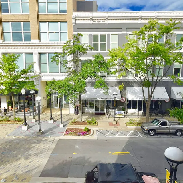 A street view of a modern urban area featuring shops with outdoor seating, trees lining the walkways, and parked cars.