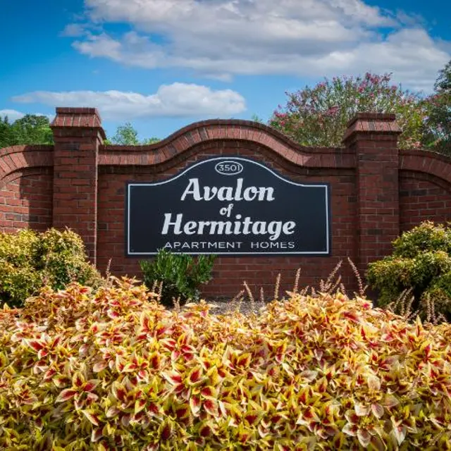 A brick sign for Avalon of Hermitage Apartment Homes, surrounded by colorful plants and flowers against a blue sky.