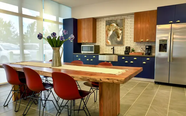 A modern kitchen and dining area featuring a large wooden dining table with black wire chairs. The kitchen has navy blue cabinets, a stainless steel refrigerator, and a large window letting in natural light. A vase with purple flowers is centered on the table.