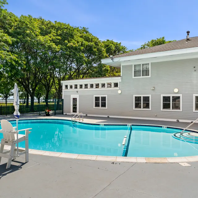 A serene swimming pool area with a light blue pool surrounded by concrete and a building in the background. Trees and a fence are visible in the background, adding to the relaxing atmosphere.