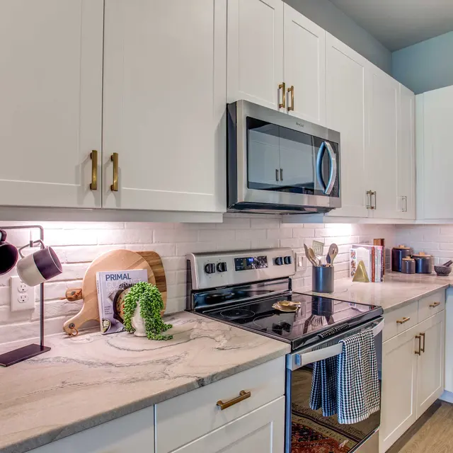 A modern kitchen with white cabinets, stainless steel appliances, and a marble countertop. There are various utensils and decorative items on the counter, along with a small plant for decoration.