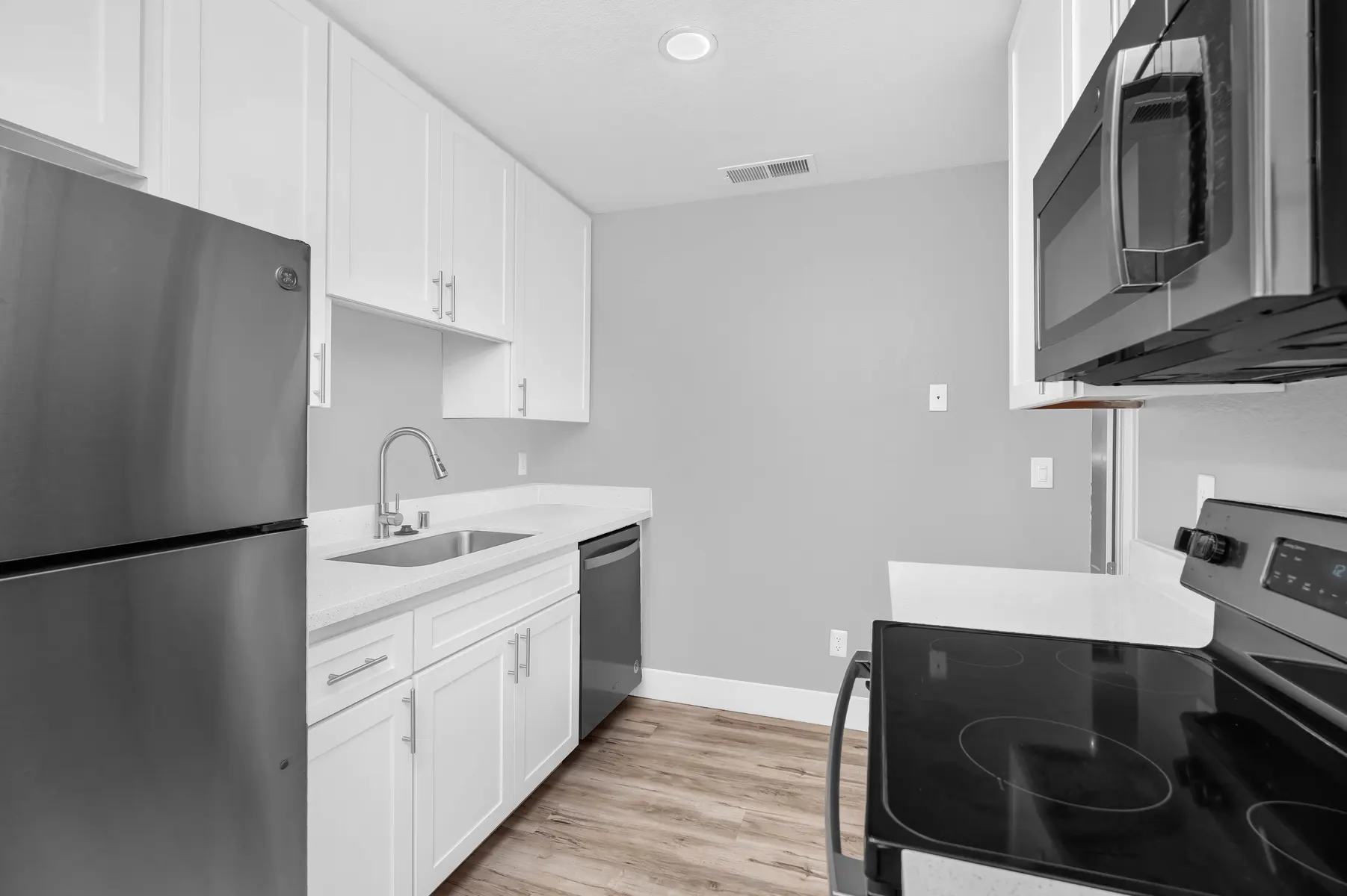 A modern kitchen featuring white cabinetry, stainless steel appliances, and light wood flooring. The kitchen includes a refrigerator, a dishwasher, and an electric stovetop, complemented by a sleek sink and minimalist design.