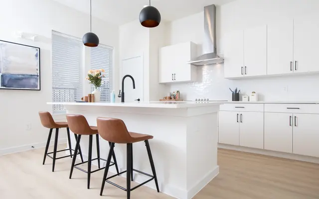 A modern kitchen featuring white cabinets, a central island with four brown bar stools, pendant lighting, and a stainless steel range hood. Soft natural light filters through a window with blinds.
