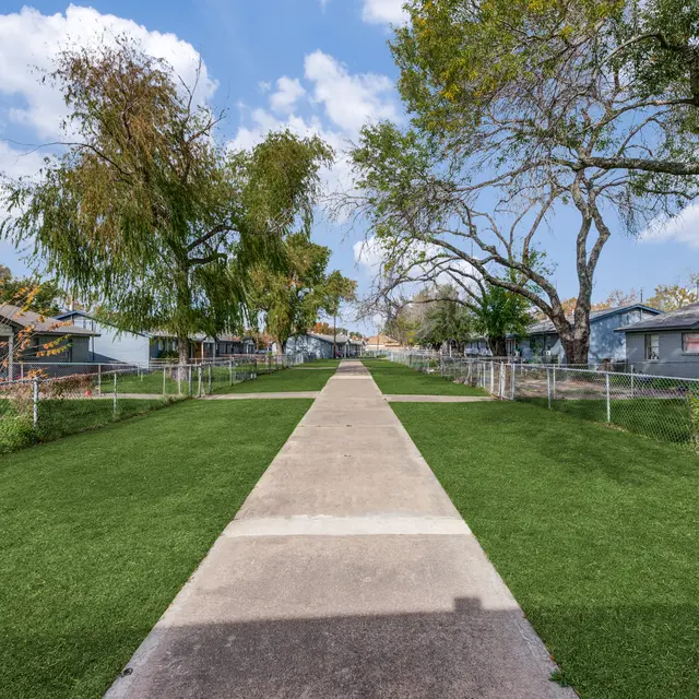 Pathway Through Residential Area A paved pathway leading through a green lawn with fenced yards on either side. Trees line the pathway under a partly cloudy sky.