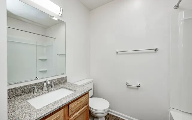 A clean, modern bathroom featuring a granite countertop, a large mirror, a white toilet, and a bathtub with a shower. The walls are painted white, and there is a towel bar and shelves for storage.