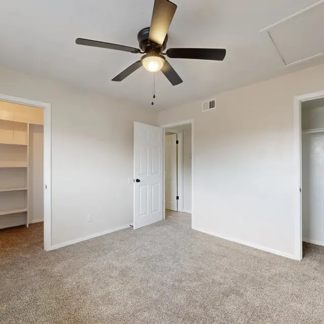 A spacious, empty bedroom featuring beige carpet, light-colored walls, and a ceiling fan. The room has two open doors leading to closets and a built-in shelf area on the left side.