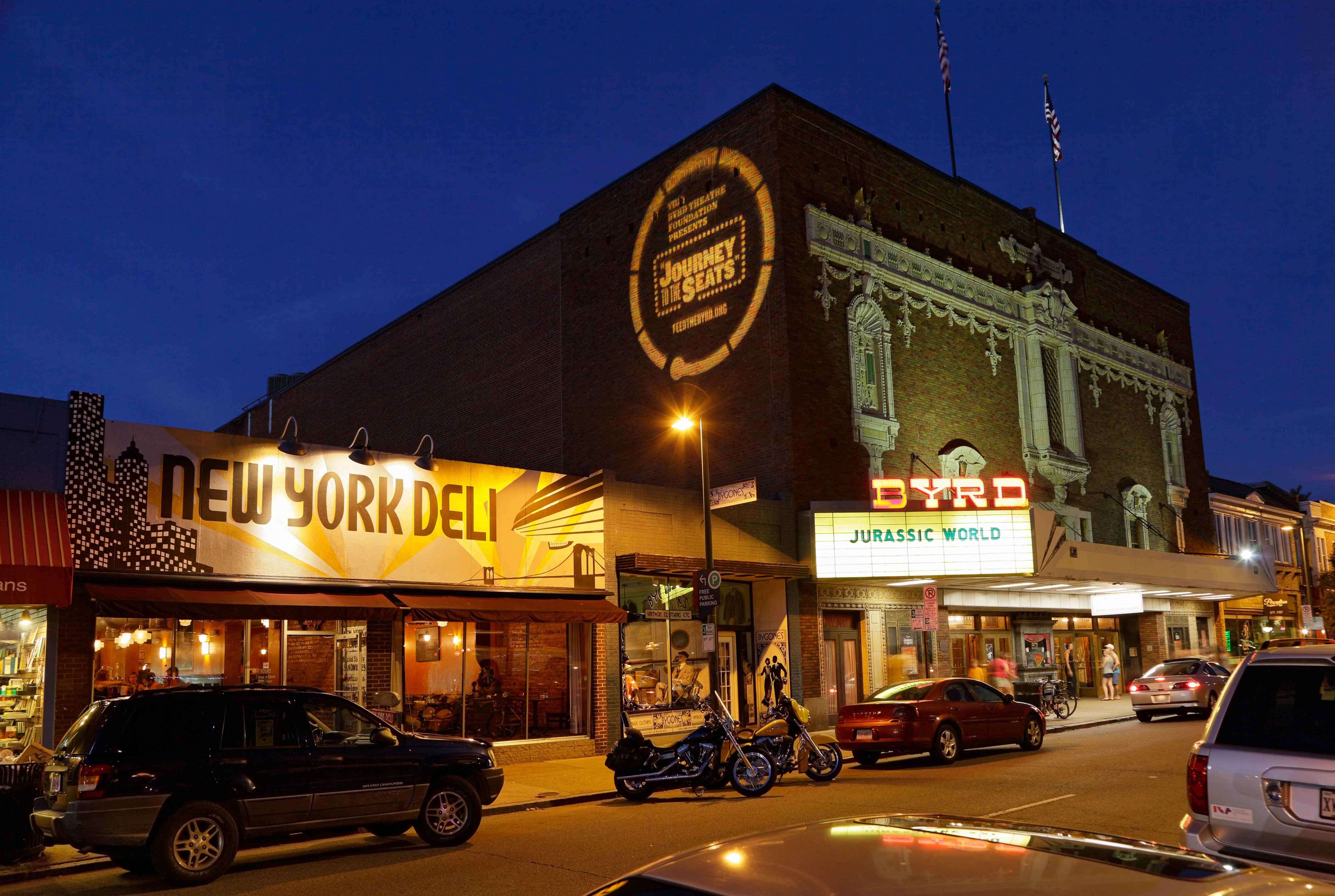 A nighttime view of a street featuring a deli and a theater. The deli has a prominent sign that reads 'NEW YORK DELI', and the theater displays a poster for 'JURASSIC WORLD'. The building is illuminated with warm lights against a dark sky.