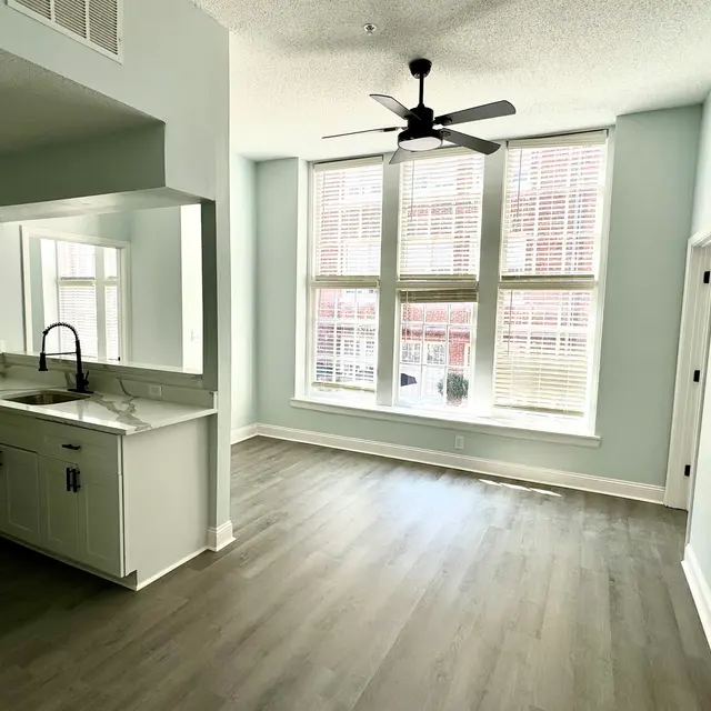 A modern apartment interior featuring light blue walls, large windows with blinds, and a ceiling fan. There is a kitchen area with a sink and cabinets visible.
