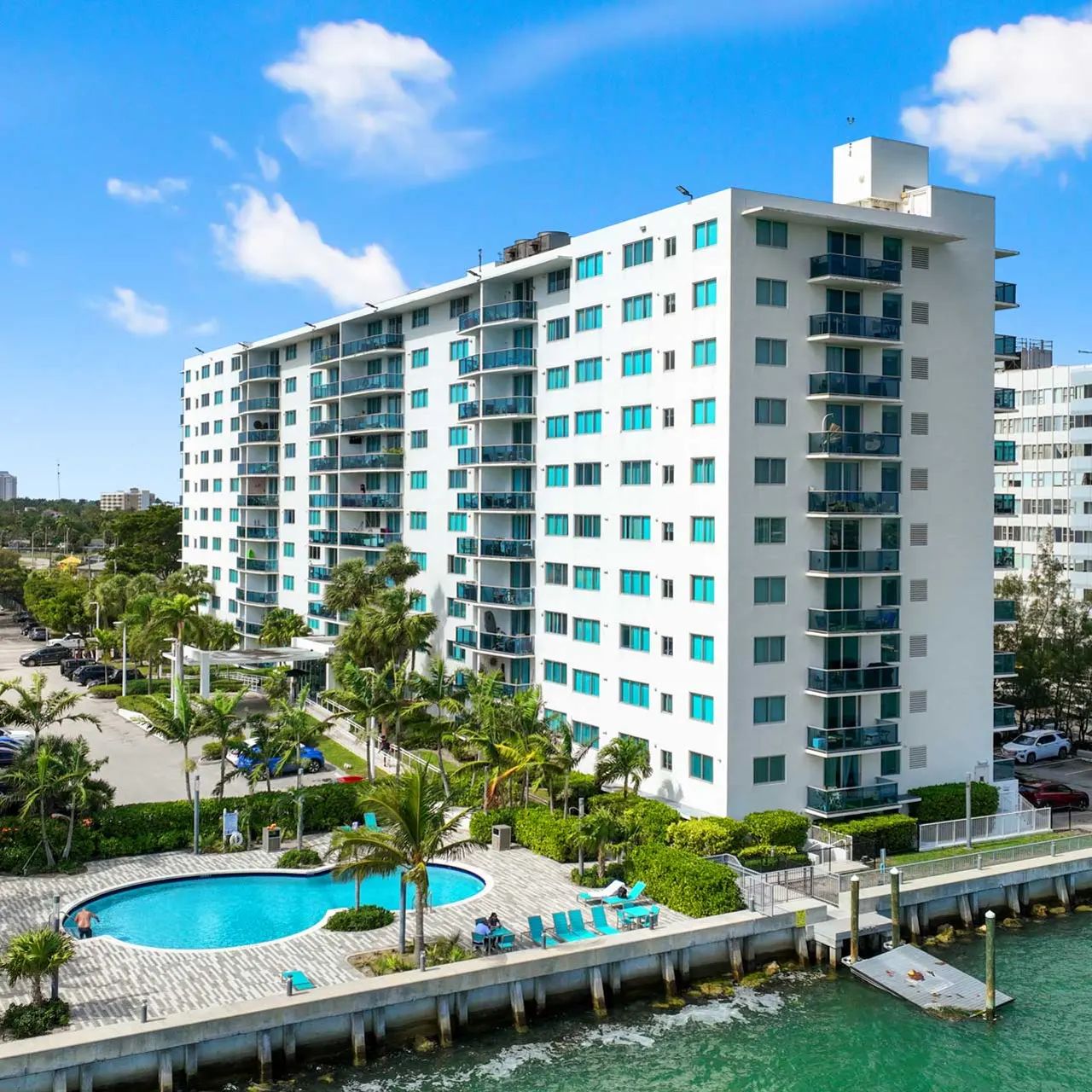 An oceanfront apartment building with a swimming pool in front, surrounded by lush greenery and parking areas.