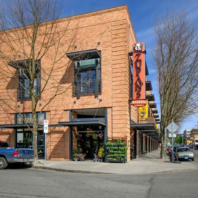 A corner building with a brick facade, featuring large windows and a sign that reads 'Zokays'. Leafless trees are visible in front, and there are parked cars on the street.