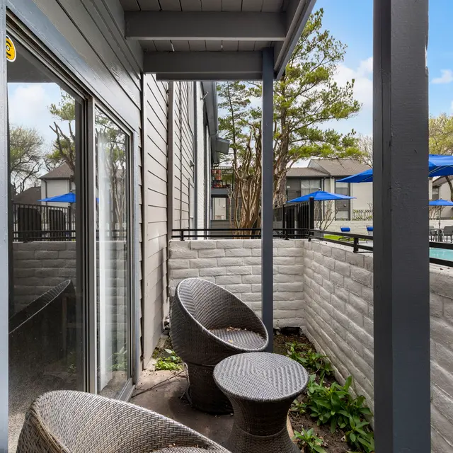 Outdoor Patio Area A small outdoor patio area with two wicker chairs and a round table, surrounded by a low wall. There are green plants at the base of the wall and a view of a swimming pool area beyond.