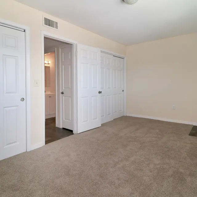 A spacious empty room featuring beige walls and carpeting, with multiple white closet doors on the left and a bathroom door visible in the background.