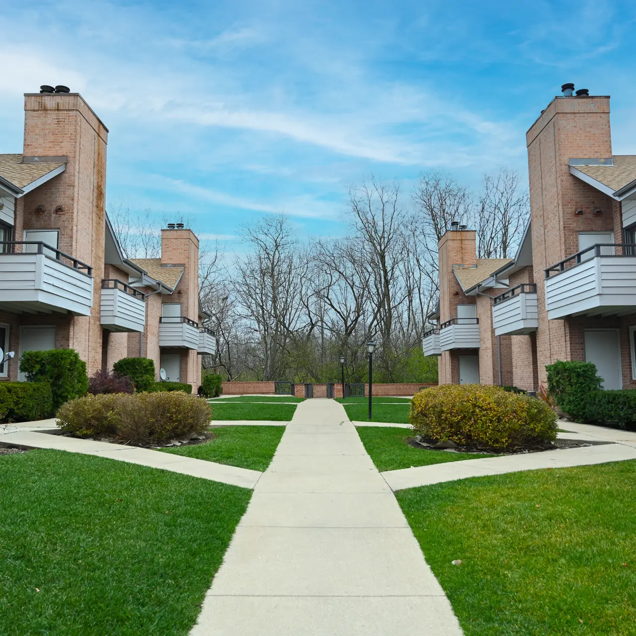 A view of a residential apartment complex featuring two rows of brick buildings with balconies, surrounded by well-maintained lawns and shrubbery. A concrete pathway leads through the center of the area, and trees are visible in the background under a blue sky.