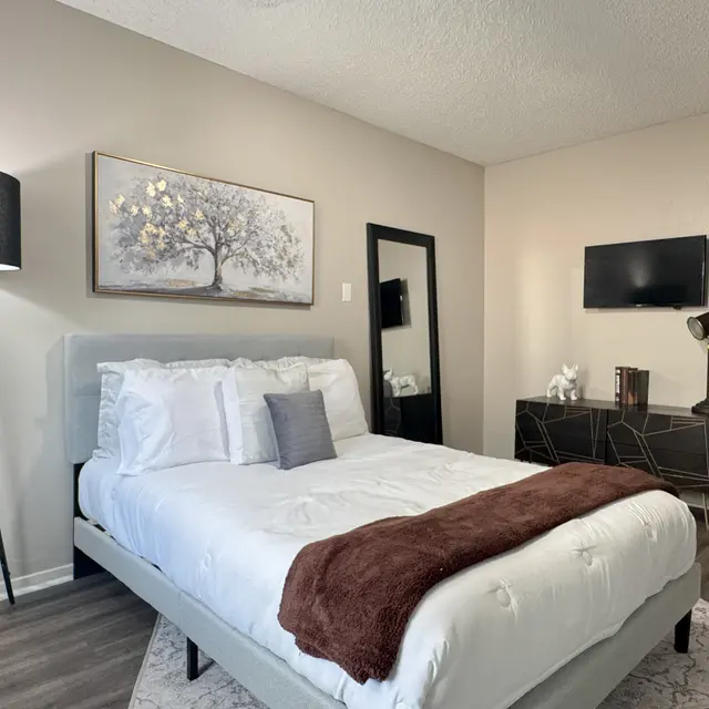 A modern bedroom featuring a bed with white bedding, a brown throw, a floor lamp, an abstract painting, a full-length mirror, a TV mounted on the wall, and contemporary decor.