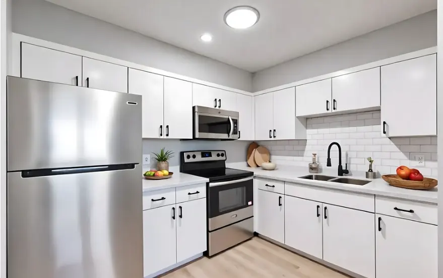 A modern kitchen featuring stainless steel appliances, white cabinets, and a light-colored countertop. The kitchen is well-lit with a round ceiling light, and there are fruits displayed on a plate on the counter.
