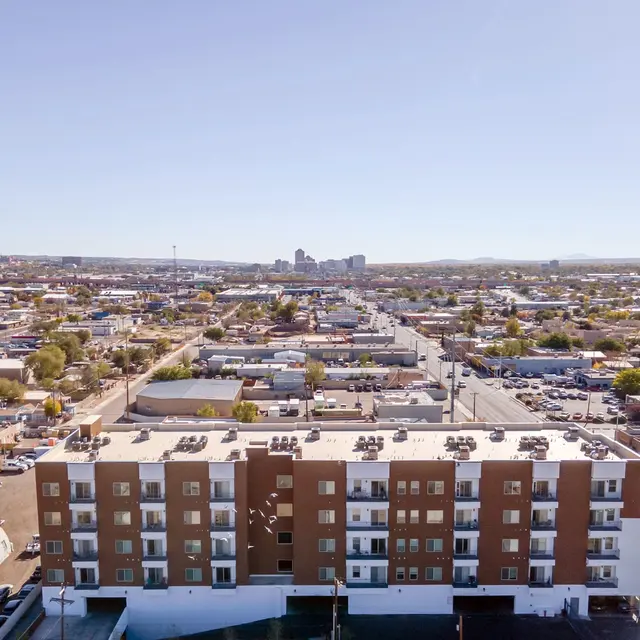 Aerial view of a cityscape showing a mix of residential and commercial buildings. In the foreground, a multi-story building with a flat roof is visible, while the background features many small houses, streets, and distant skyline.