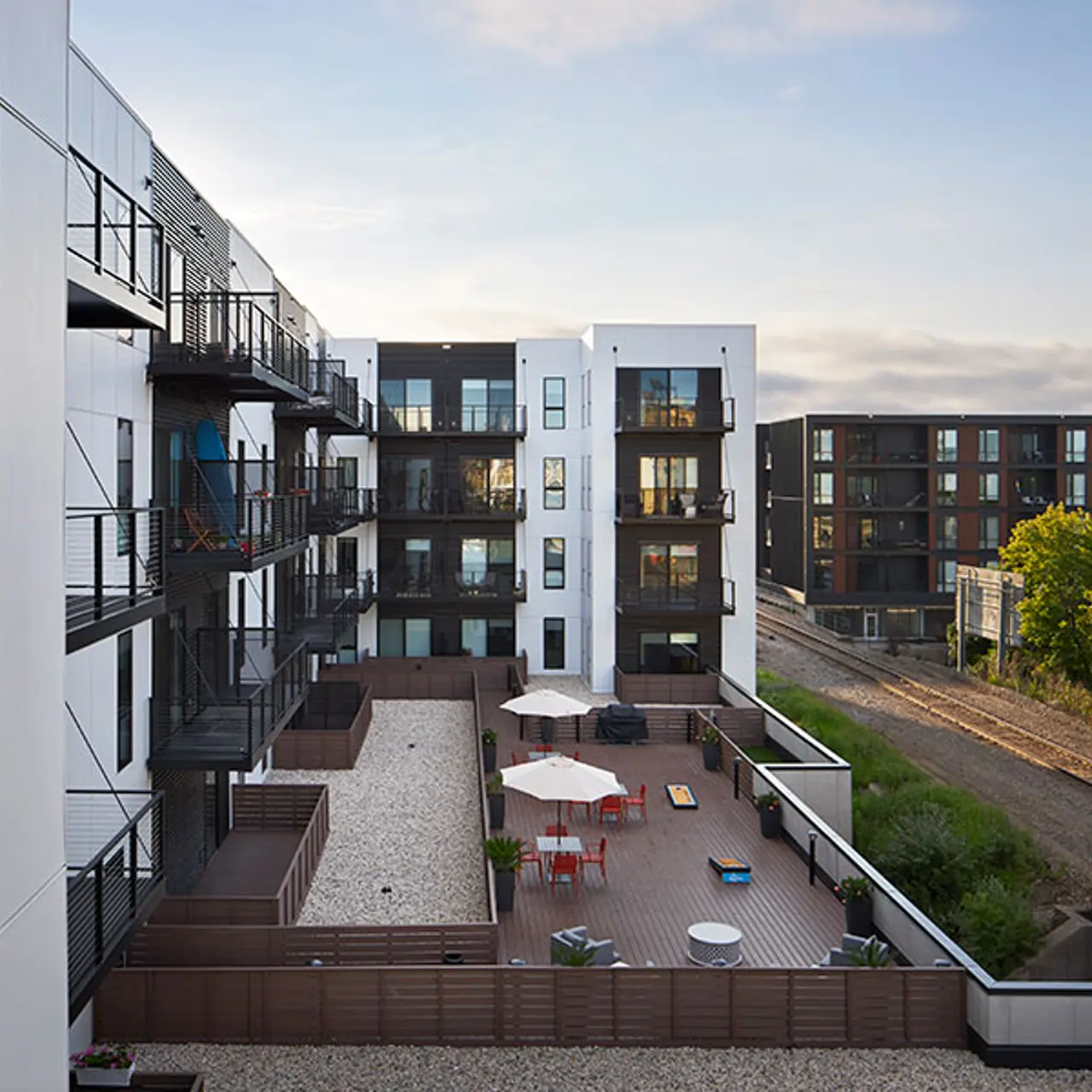 Modern Apartment Complex with Outdoor Amenities A contemporary apartment complex with multiple balconies and a shared outdoor space featuring patio furniture and umbrellas. The setting includes green trees and a view of a nearby railway.