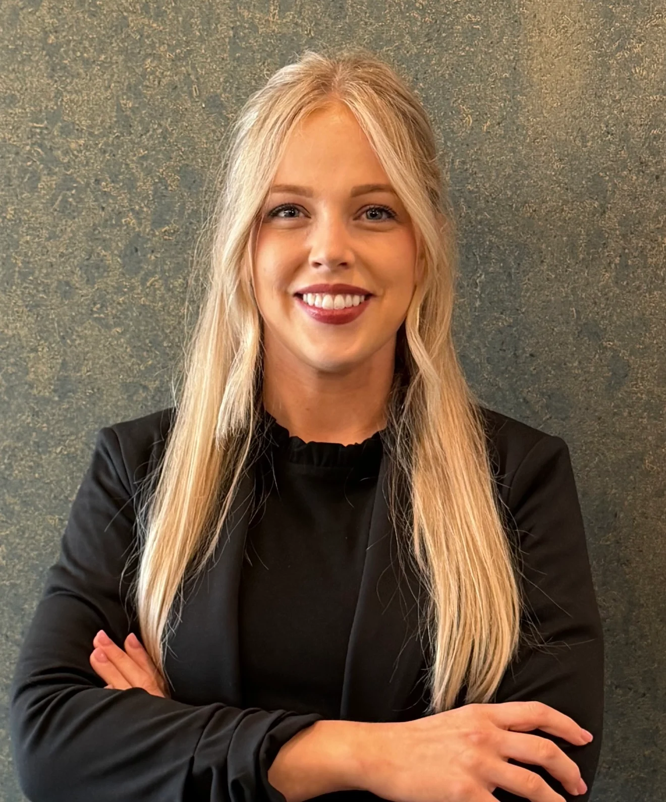 Professional Portrait A woman with long blonde hair styled straight, wearing a black blazer, smiling at the camera with her arms crossed.
